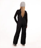 A model shown from the back wearing a black long-sleeve top and black snow bibs, paired with a gray knit beanie and gray boots, against a solid white studio background.
