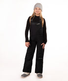 A young girl modeling black snow bib overalls with a gray beanie, black long-sleeve shirt, and gray boots against a white studio background.