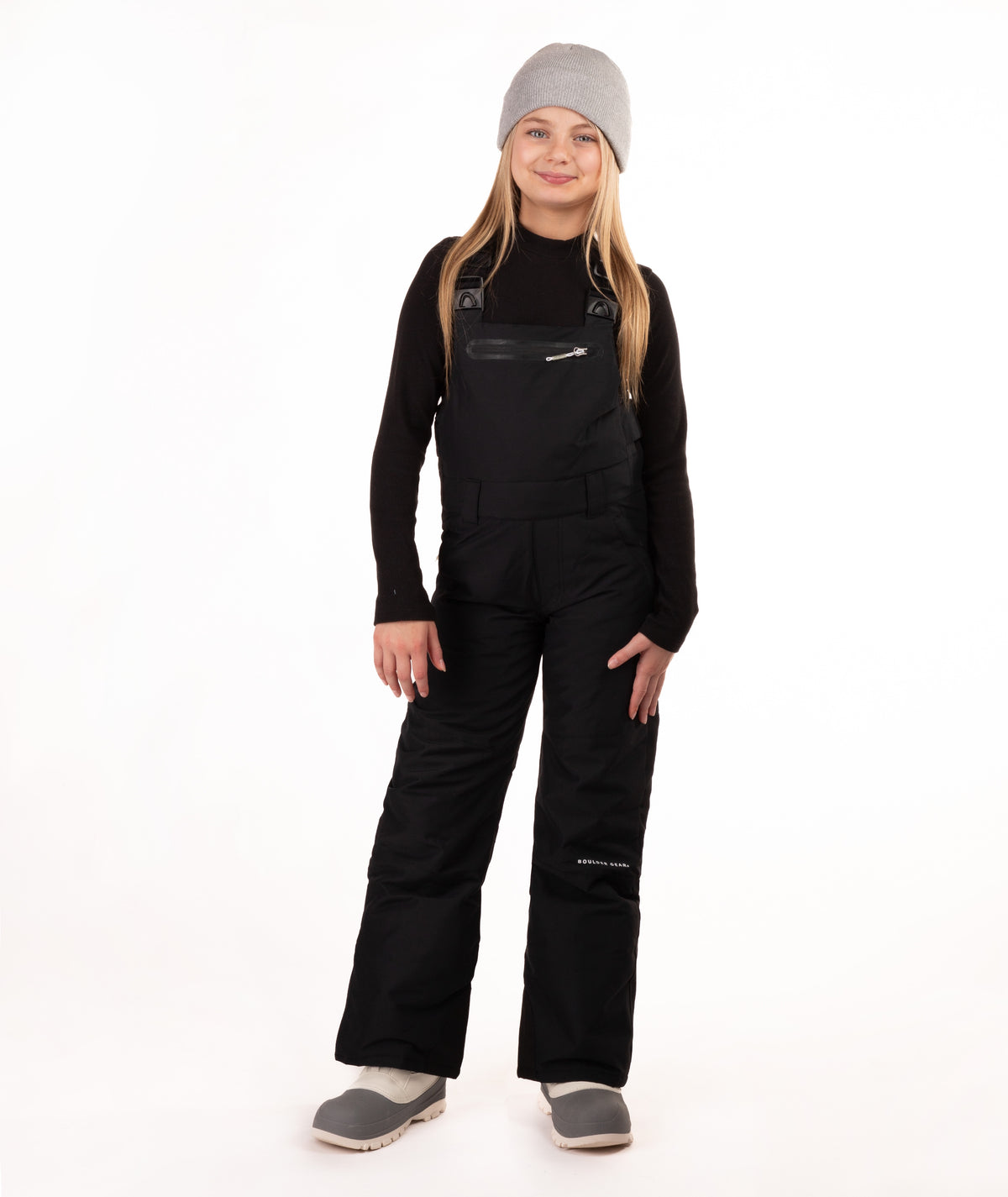 A young girl modeling black snow bib overalls with a gray beanie, black long-sleeve shirt, and gray boots against a white studio background.