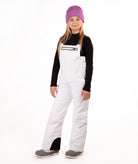 A young girl modeling white snow bib overalls, a black long-sleeve shirt, gray boots, and a purple knit beanie, standing against a solid white background.