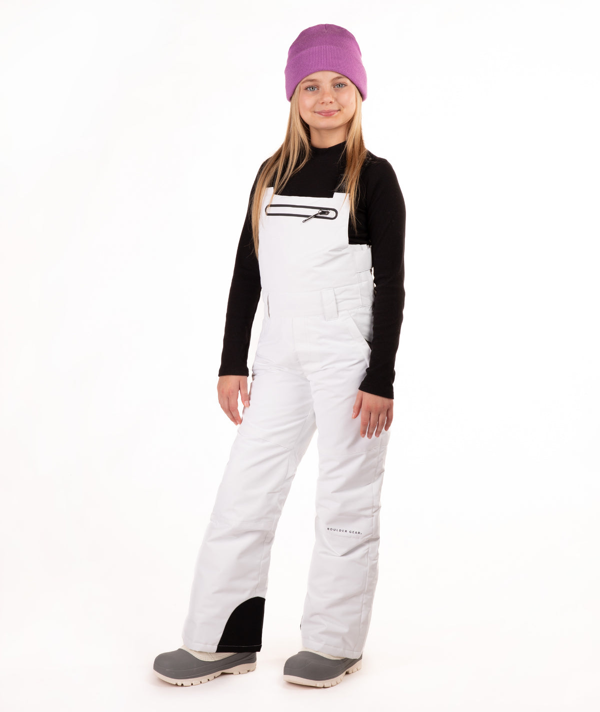 A young girl modeling white snow bib overalls, a black long-sleeve shirt, gray boots, and a purple knit beanie, standing against a solid white background.