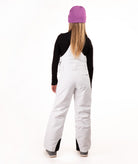 A model viewed from the back wearing white snow bibs, a black long-sleeve top, a purple beanie, and gray boots, set against a plain white studio background.
