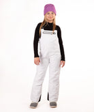 A young girl modeling white snow bib overalls, a black long-sleeve shirt, gray boots, and a purple knit beanie, standing against a solid white background.