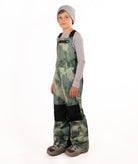 A child wearing camouflage snow bib overalls with black knee panels, a gray beanie, a light gray long-sleeve shirt, and boots, standing against a white studio background.