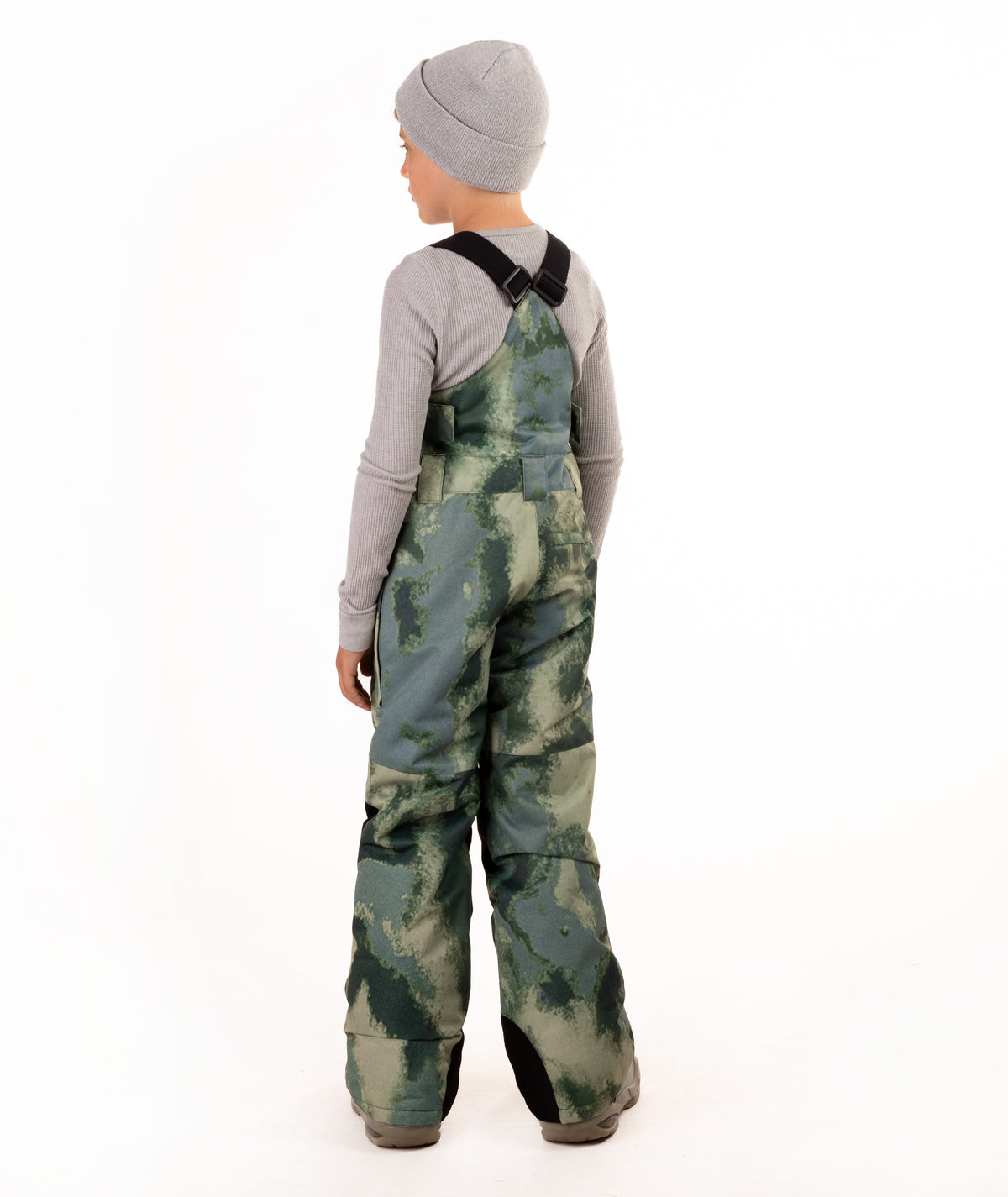 A child shown from the back wearing green and gray camouflage snow bib overalls with a gray beanie and gray long-sleeve shirt, photographed against a plain white studio background.