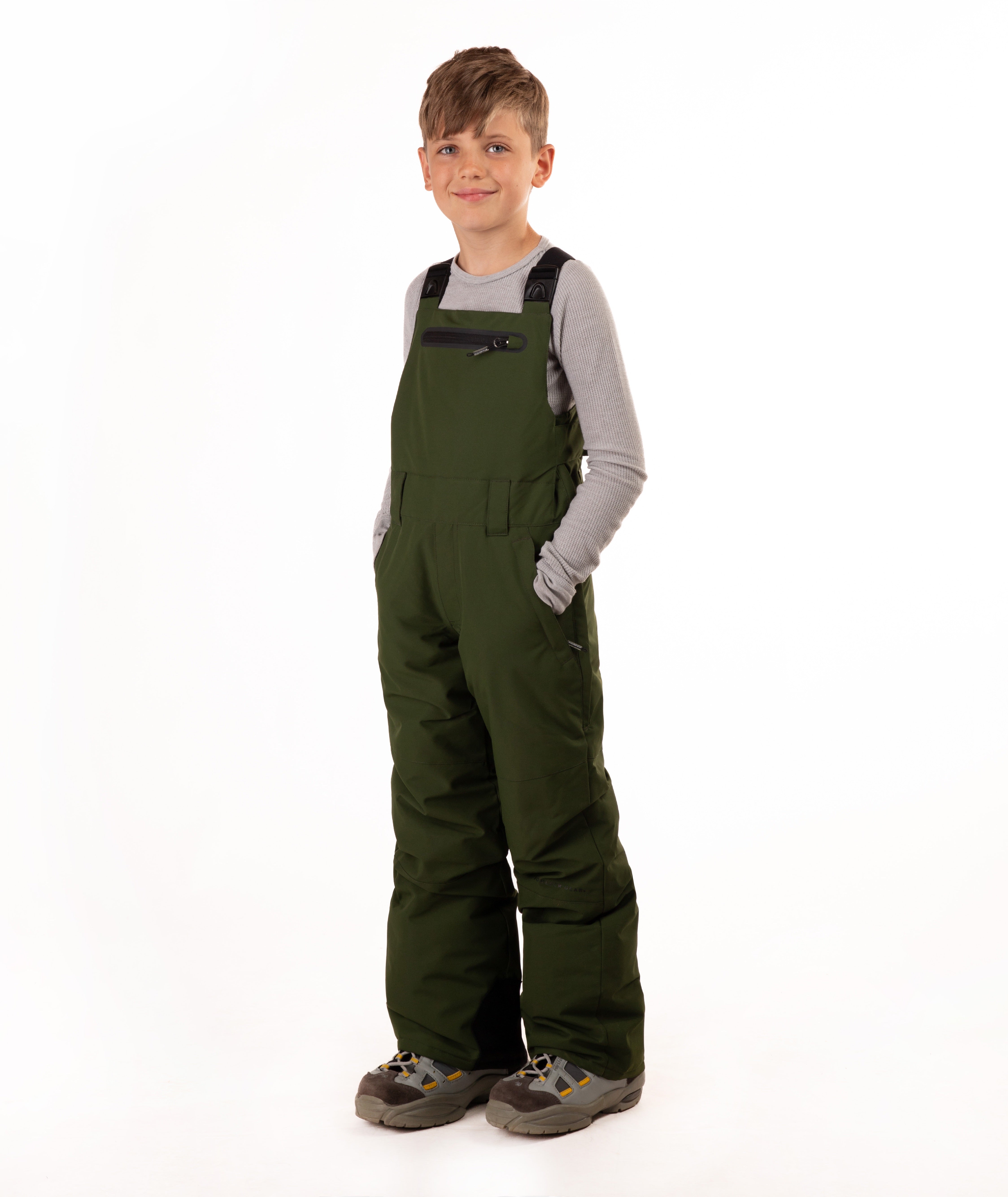 A young boy wearing dark green snow bib overalls over a gray long-sleeve shirt, standing against a solid white studio background.
