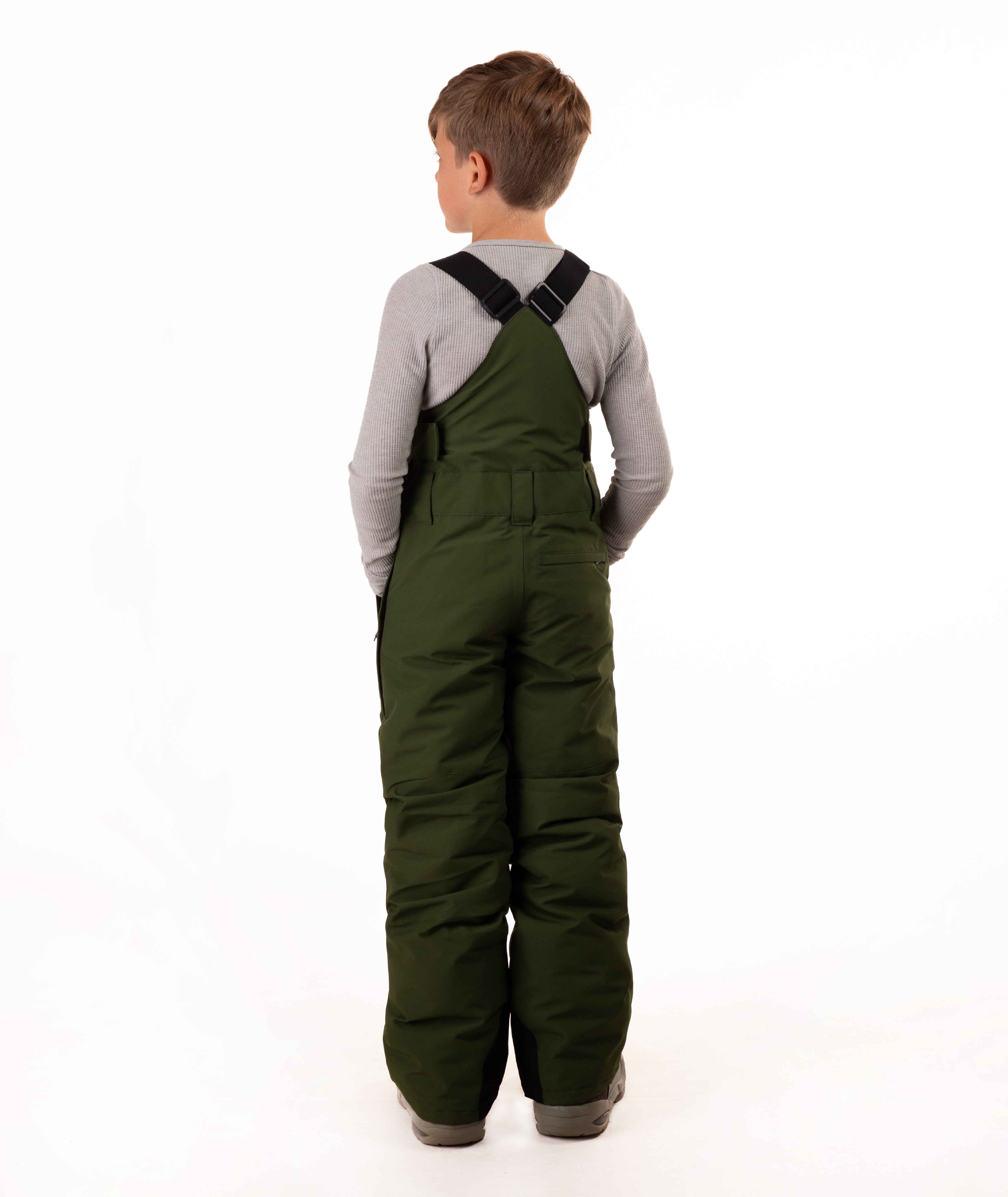 A child viewed from the back wearing dark olive green insulated bib overalls  over a light gray long-sleeve shirt, standing against a plain white studio background.