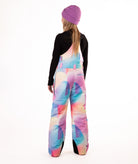back of girl wearing multi colored abstract pastel pattern snow bibs with a black top on a white background
