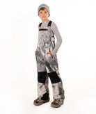 A child wearing gray and black camouflage snow bib overalls with black knee patches, a gray long-sleeve shirt, a gray beanie, and winter boots, standing against a white studio background.