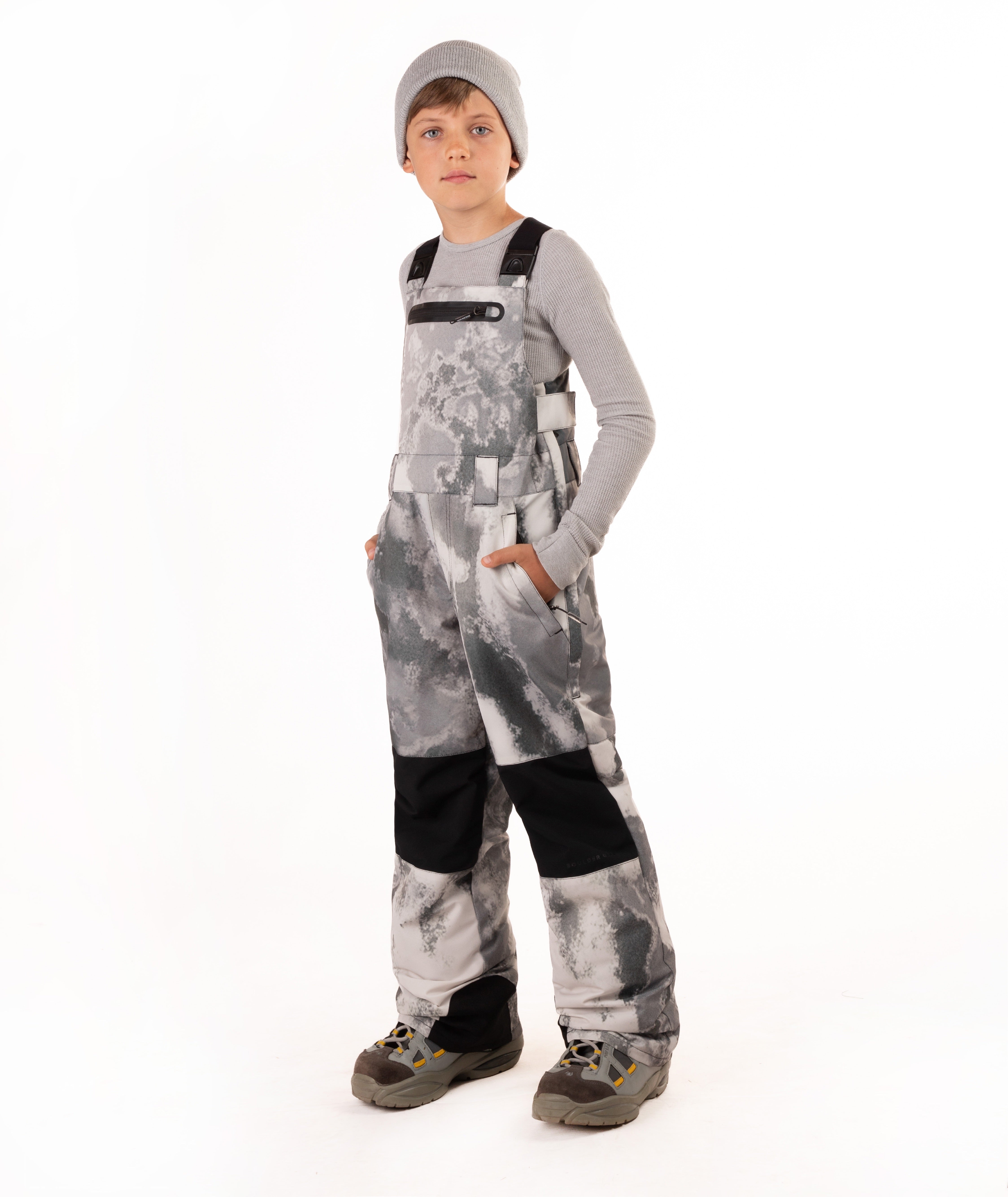 A child wearing gray and black camouflage snow bib overalls with black knee patches, a gray long-sleeve shirt, a gray beanie, and winter boots, standing against a white studio background.