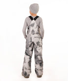 A child viewed from the back wearing gray and white tie-dye snow bib overalls, a gray long-sleeve shirt, and a gray beanie on a white studio background.