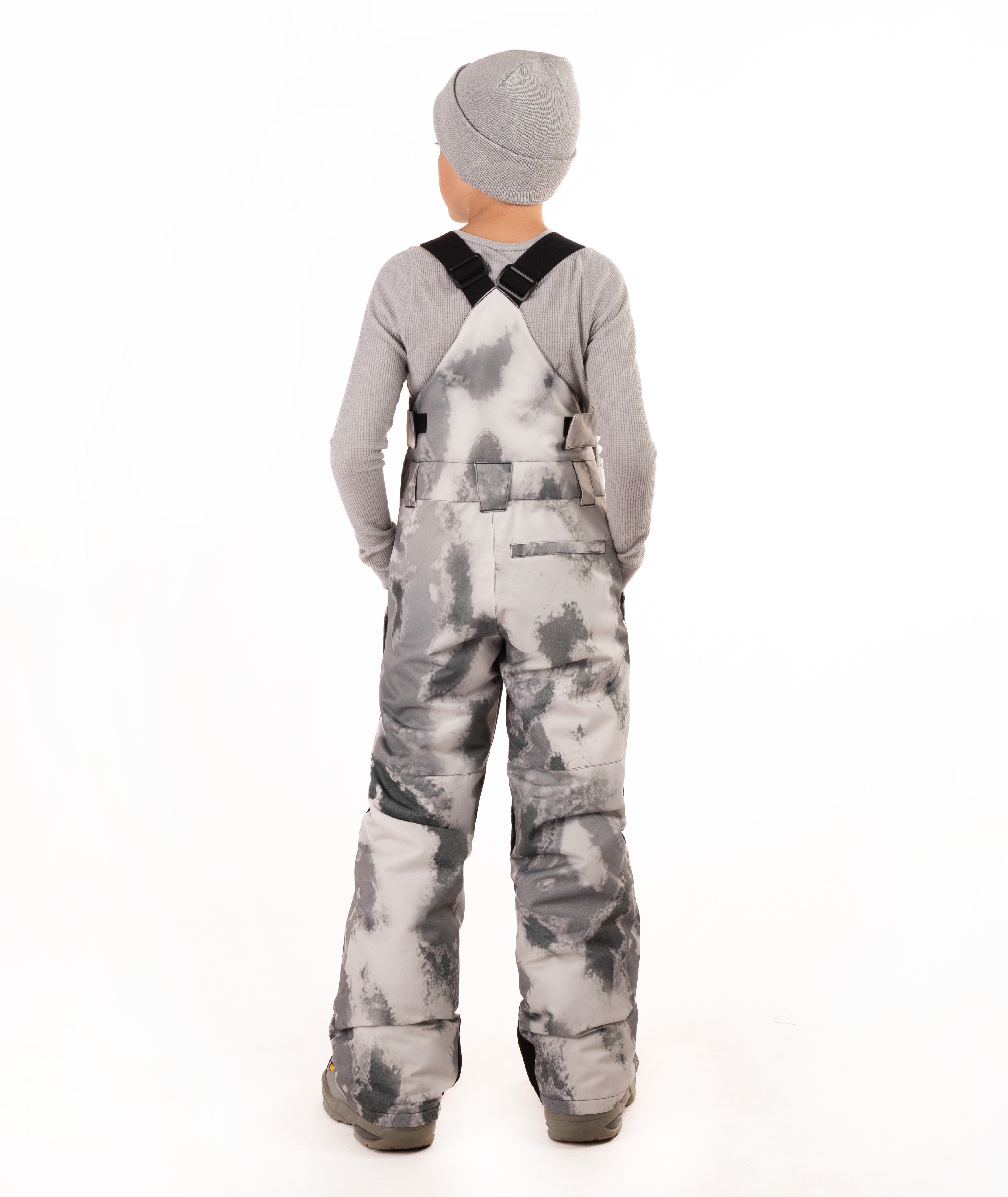 A child viewed from the back wearing gray and white tie-dye snow bib overalls, a gray long-sleeve shirt, and a gray beanie on a white studio background.