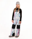 A young girl wearing a black long-sleeve shirt and colorful snow bib overalls with a white base and purple, blue, and pink paint-stroke pattern, along with a gray beanie and gray shoes, standing against a white studio background.