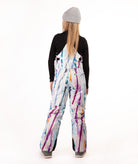 A model viewed from behind wearing a gray beanie, a black long-sleeve shirt, and white snow bibs with a colorful paint-splatter pattern in purple, blue, orange, and yellow, set against a plain white studio background.