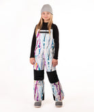 A young girl wearing a black long-sleeve shirt and colorful snow bib overalls with a white base and purple, blue, and pink paint-stroke pattern, along with a gray beanie and gray shoes, standing against a white studio background.