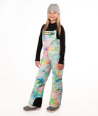 A girl wearing a gray beanie, black long-sleeve shirt, and colorful pastel tie-dye snow bib overalls with gray boots, standing against a plain white studio background.