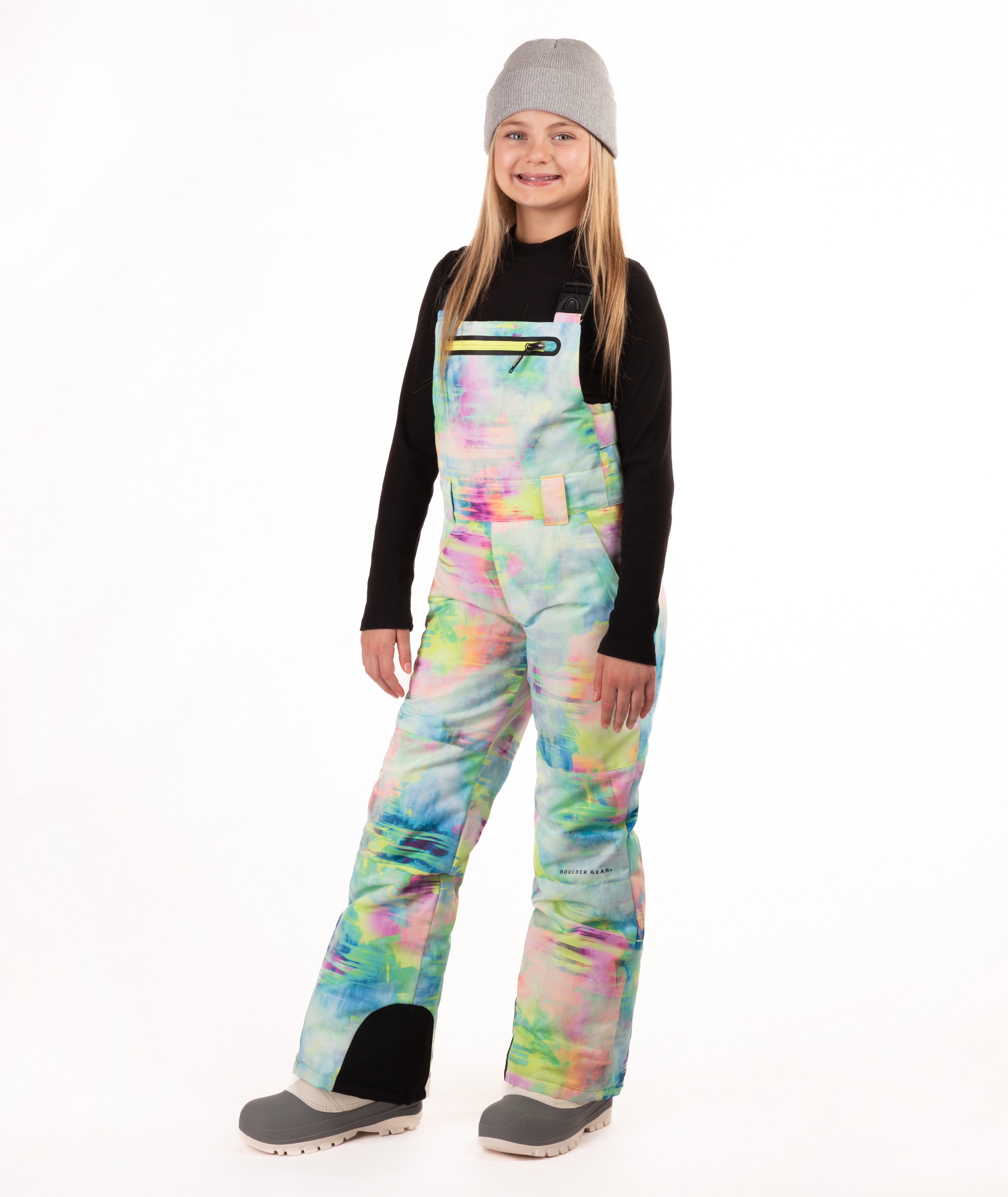 A girl wearing a gray beanie, black long-sleeve shirt, and colorful pastel tie-dye snow bib overalls with gray boots, standing against a plain white studio background.