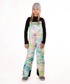 A girl wearing a gray beanie, black long-sleeve shirt, and colorful pastel tie-dye snow bib overalls with gray boots, standing against a plain white studio background.