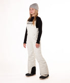 girl wearing cream colored snow bibs with a black top on a white background