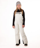 girl wearing cream colored snow bibs with a black top on a white background