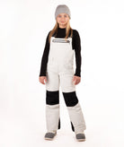 A child model wearing off-white snow bibs with black knee panels, a black long-sleeve shirt, a gray beanie, and gray boots, standing against a plain white studio background.