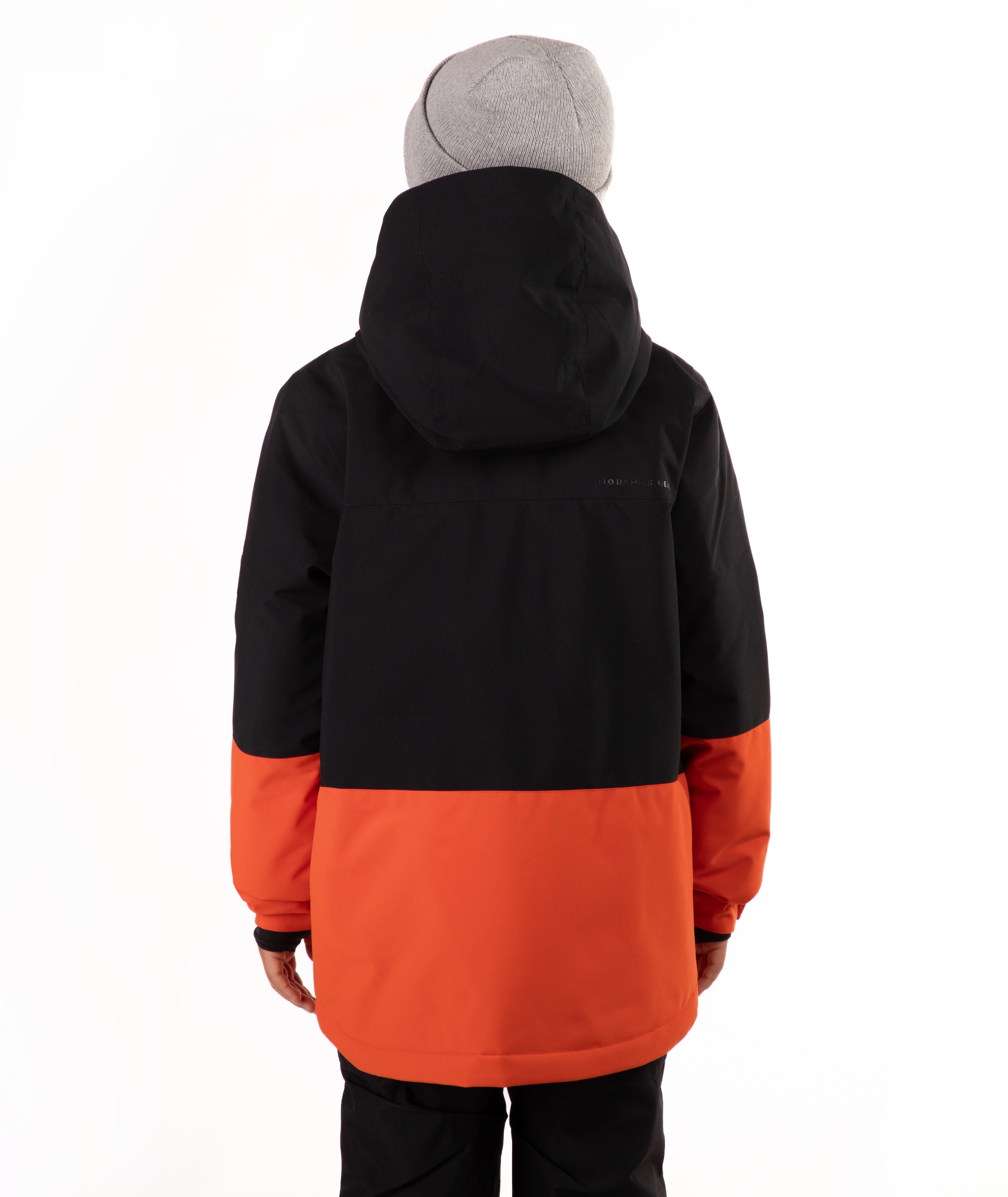 A child wearing a black and orange color-blocked hooded snow jacket, a gray knit beanie, and black pants, standing against a plain white studio background.