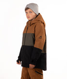 A child wearing a color-blocked ski jacket in brown, green, and black, paired with brown pants and a gray beanie, photographed against a solid white background.