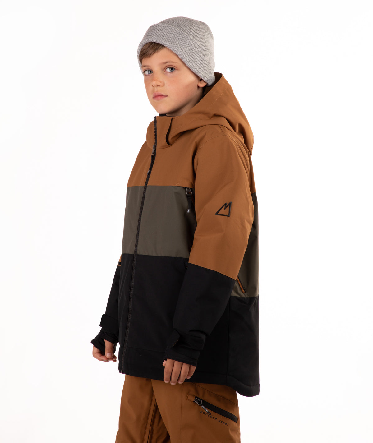A child wearing a color-blocked ski jacket in brown, green, and black, paired with brown pants and a gray beanie, photographed against a solid white background.