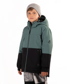 A child wearing a color-blocked ski jacket in sage green and black, paired with a gray beanie and patterned ski pants, standing against a plain white background.