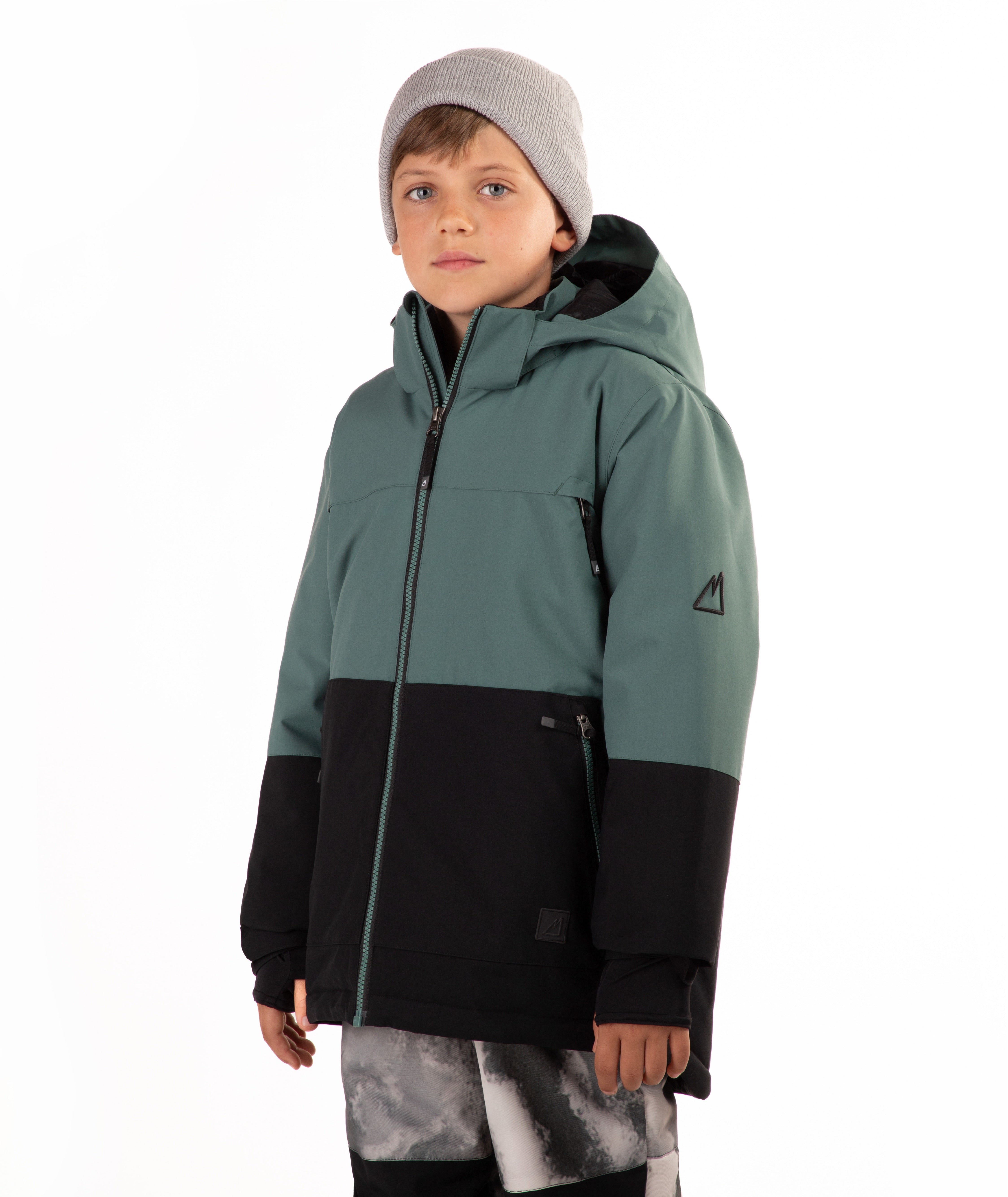 A child wearing a color-blocked ski jacket in sage green and black, paired with a gray beanie and patterned ski pants, standing against a plain white background.