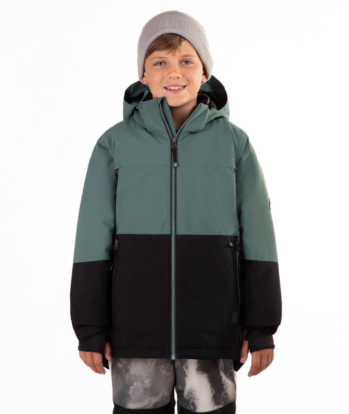 A child wearing a color-blocked ski jacket in sage green and black, paired with a gray beanie and patterned ski pants, standing against a plain white background.