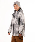 A child wearing a gray and white tie-dye patterned snowboard jacket, a gray beanie, and brown pants, standing against a plain white background.