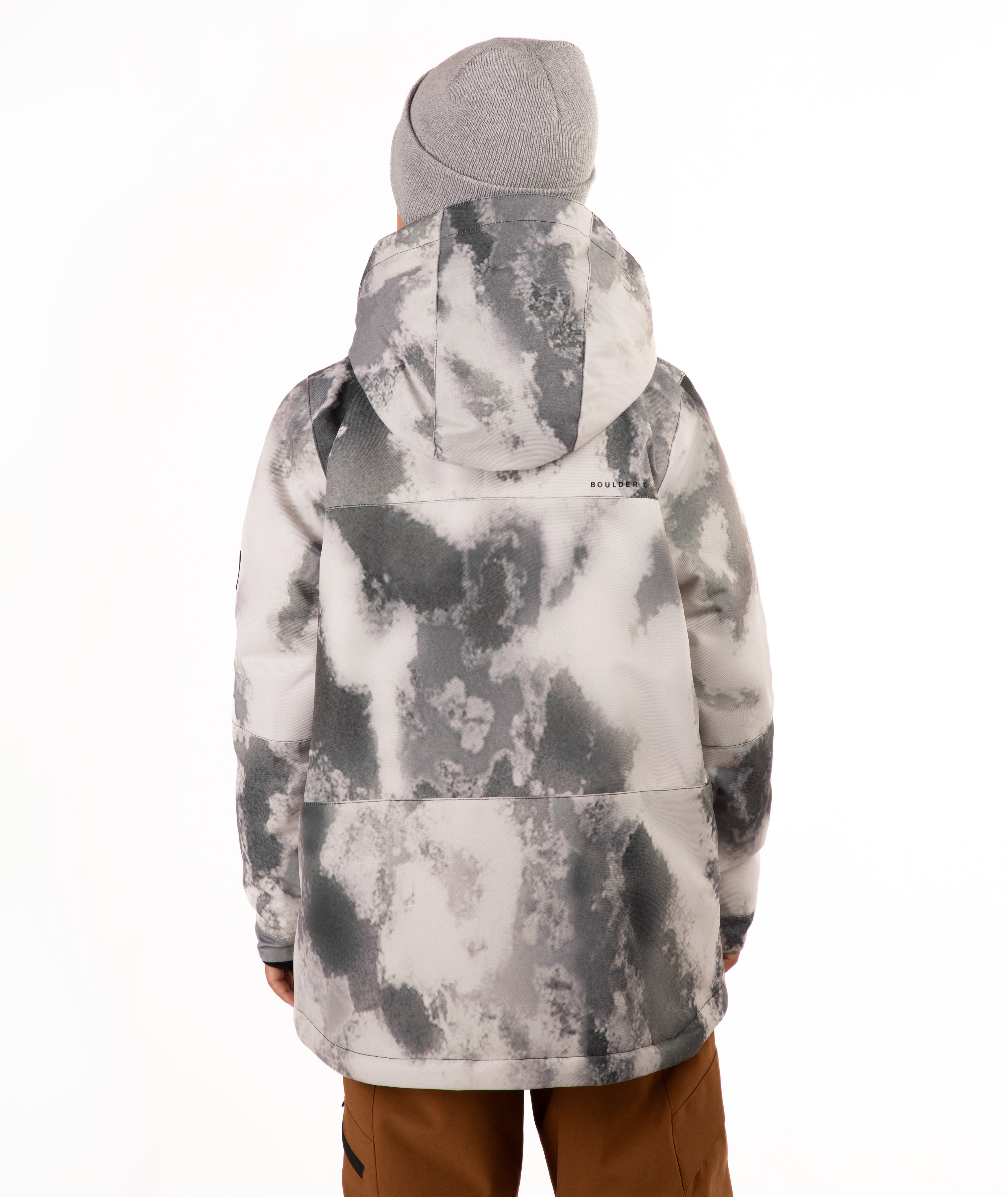 A child wearing a gray and white tie-dye patterned snowboard jacket, a gray beanie, and brown pants, standing against a plain white background.