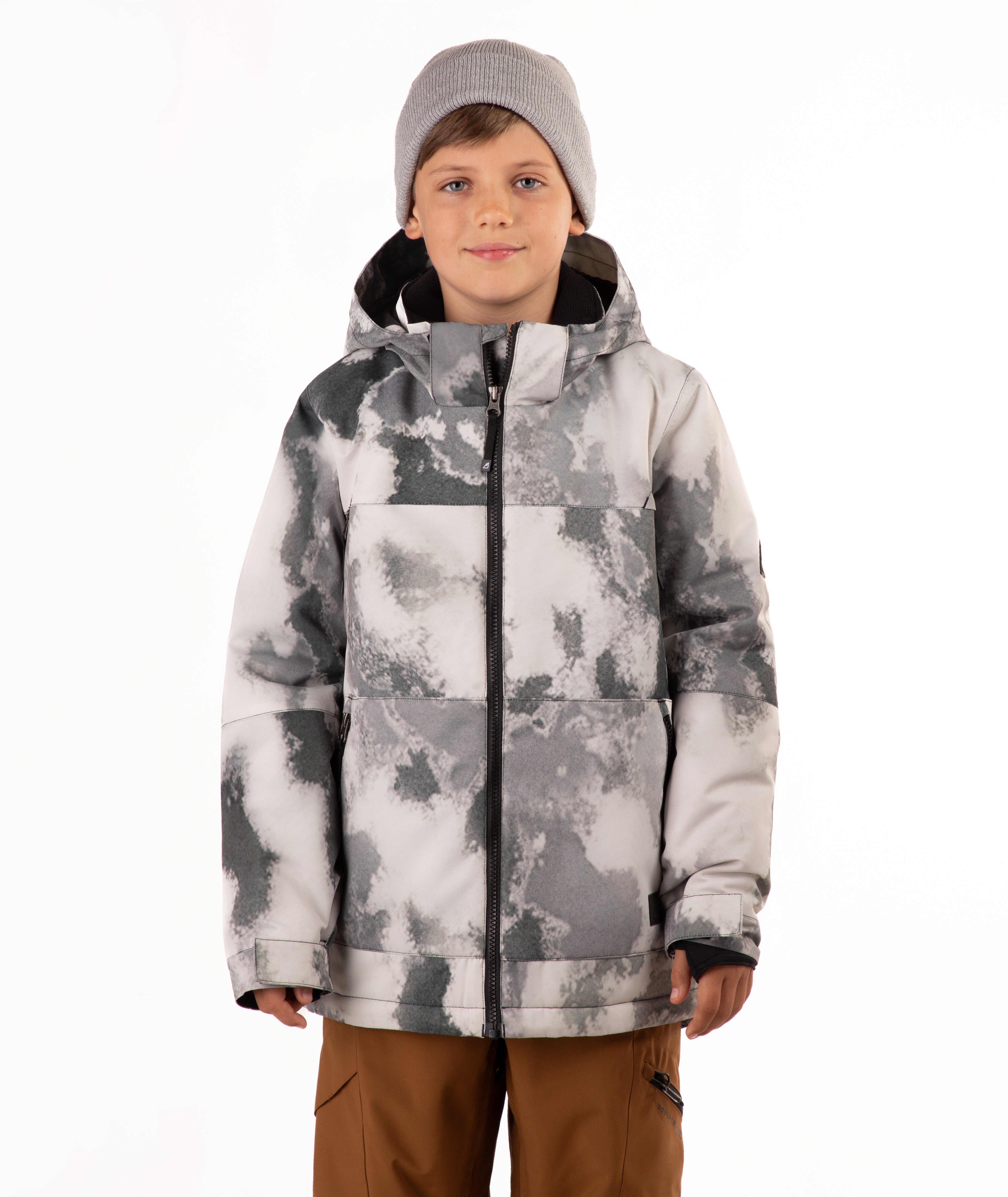 A child wearing a gray and white tie-dye patterned snowboard jacket, a gray beanie, and brown pants, standing against a plain white background.