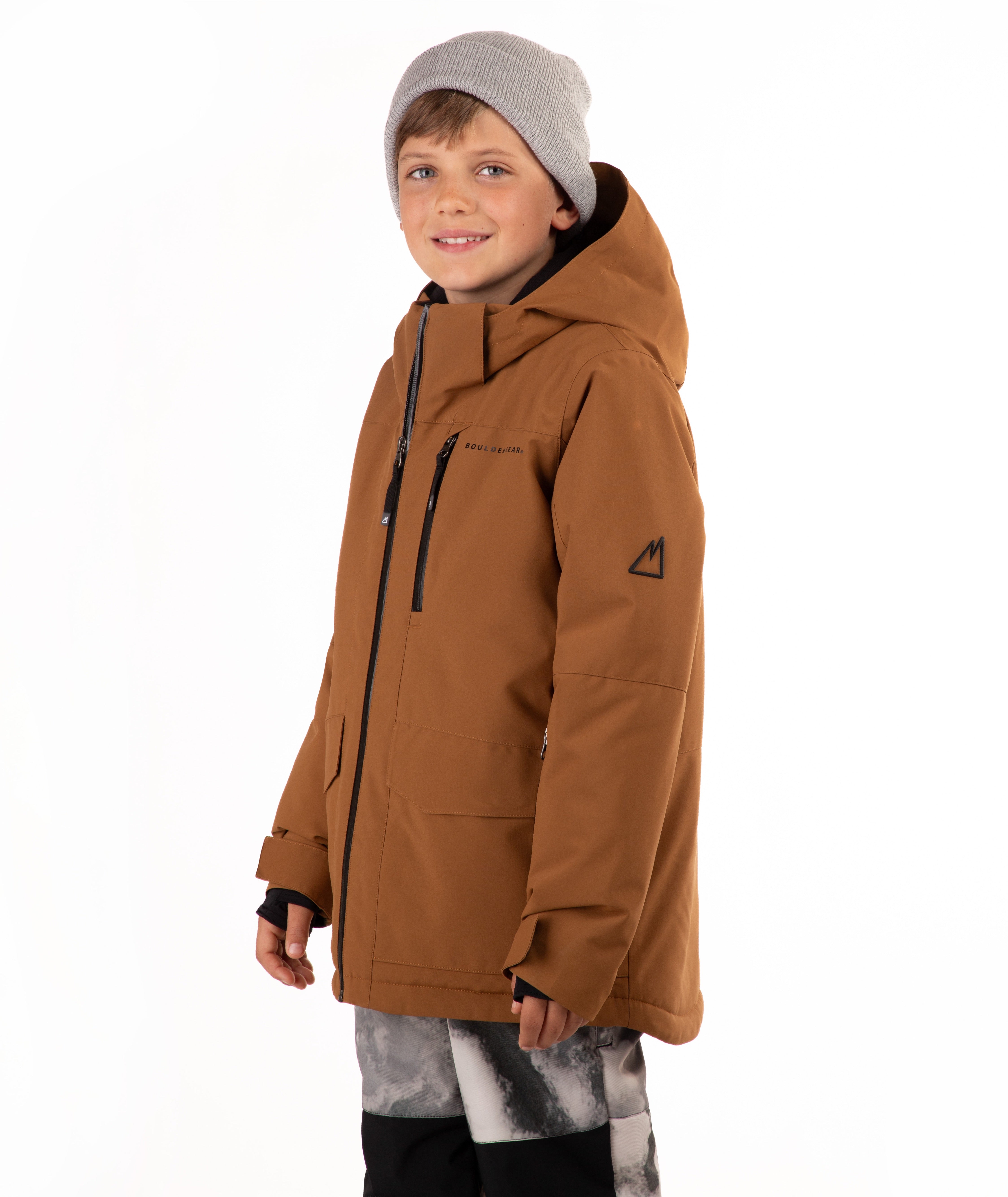 A child wearing a brown hooded snowboard jacket with black zippers, a gray beanie, and patterned pants, standing against a plain white background.
