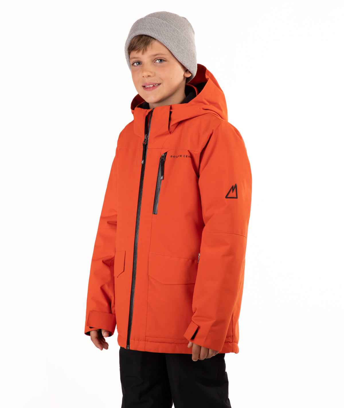A young boy wearing a bright orange hooded snowboard jacket, a gray knit beanie, and black pants, standing against a plain white studio background.