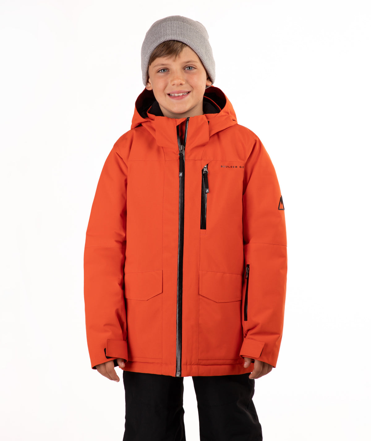A young boy wearing a bright orange hooded snowboard jacket, a gray knit beanie, and black pants, standing against a plain white studio background.
