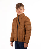 A young boy wearing a brown puffer jacket with black zippers and blue jeans, standing against a plain white background.