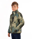 A young boy wearing a quilted puffer jacket with a green and beige camouflage pattern, standing against a plain white background.