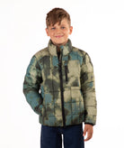 A young boy wearing a quilted puffer jacket with a green and beige camouflage pattern, standing against a plain white background.