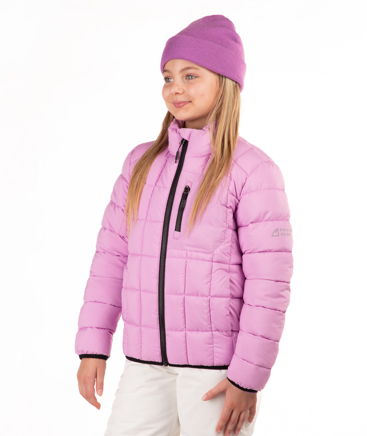 A young girl wearing a light purple puffer jacket, a matching pink knit beanie, and white pants, standing against a plain white background.