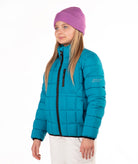 A young girl wearing a turquoise puffer jacket, a purple knit beanie, and white pants, standing against a plain white background.