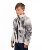 A young boy wearing a gray and white abstract patterned puffer jacket with a black front zipper, paired with blue jeans, photographed against a solid white background.