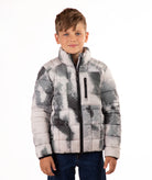 A young boy wearing a gray and white abstract patterned puffer jacket with a high collar and black zipper, paired with dark blue jeans, photographed against a solid white background.