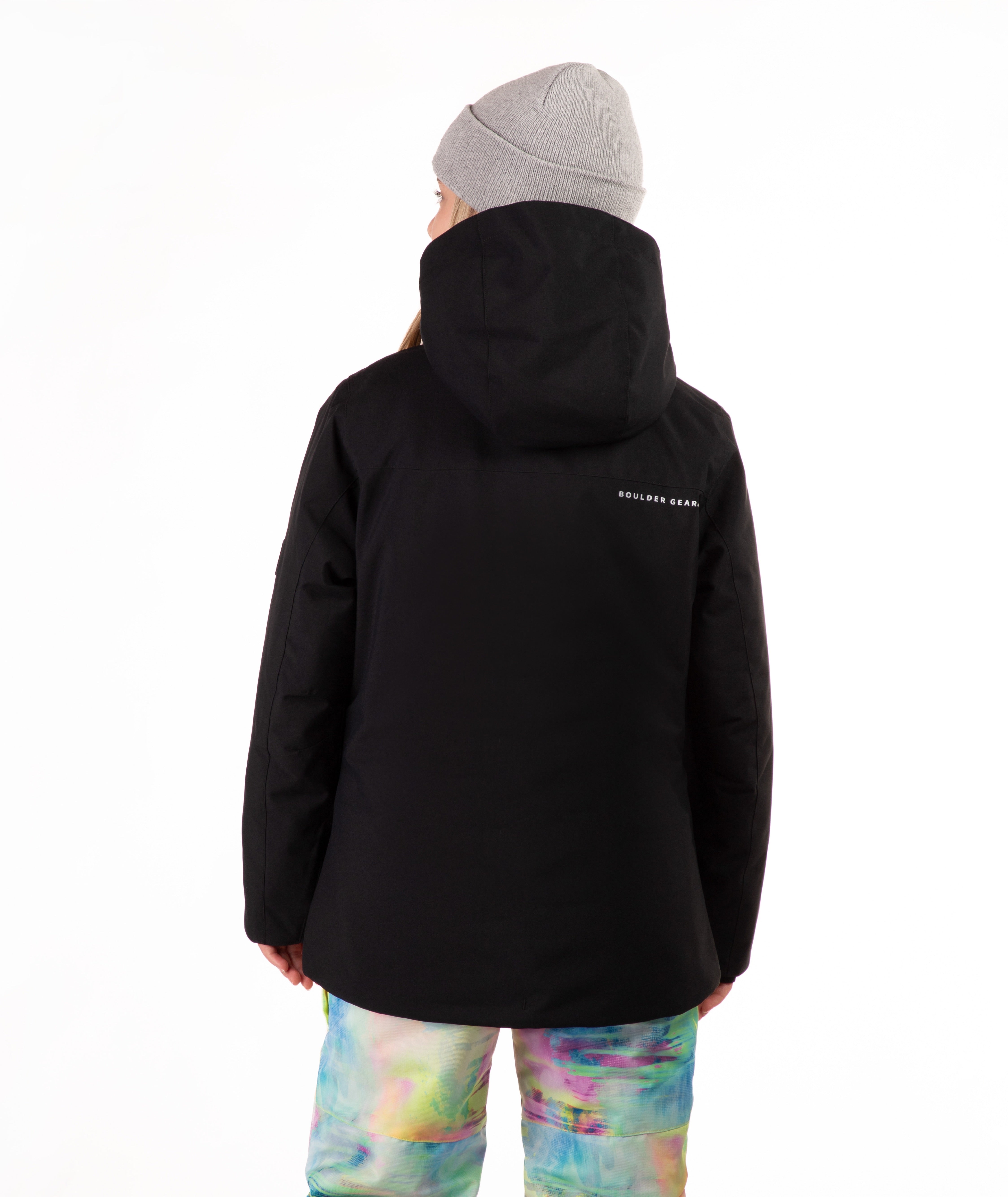 A person shown from the back wearing a black hooded jacket, a light gray beanie, and colorful tie-dye snow pants against a plain white studio background.