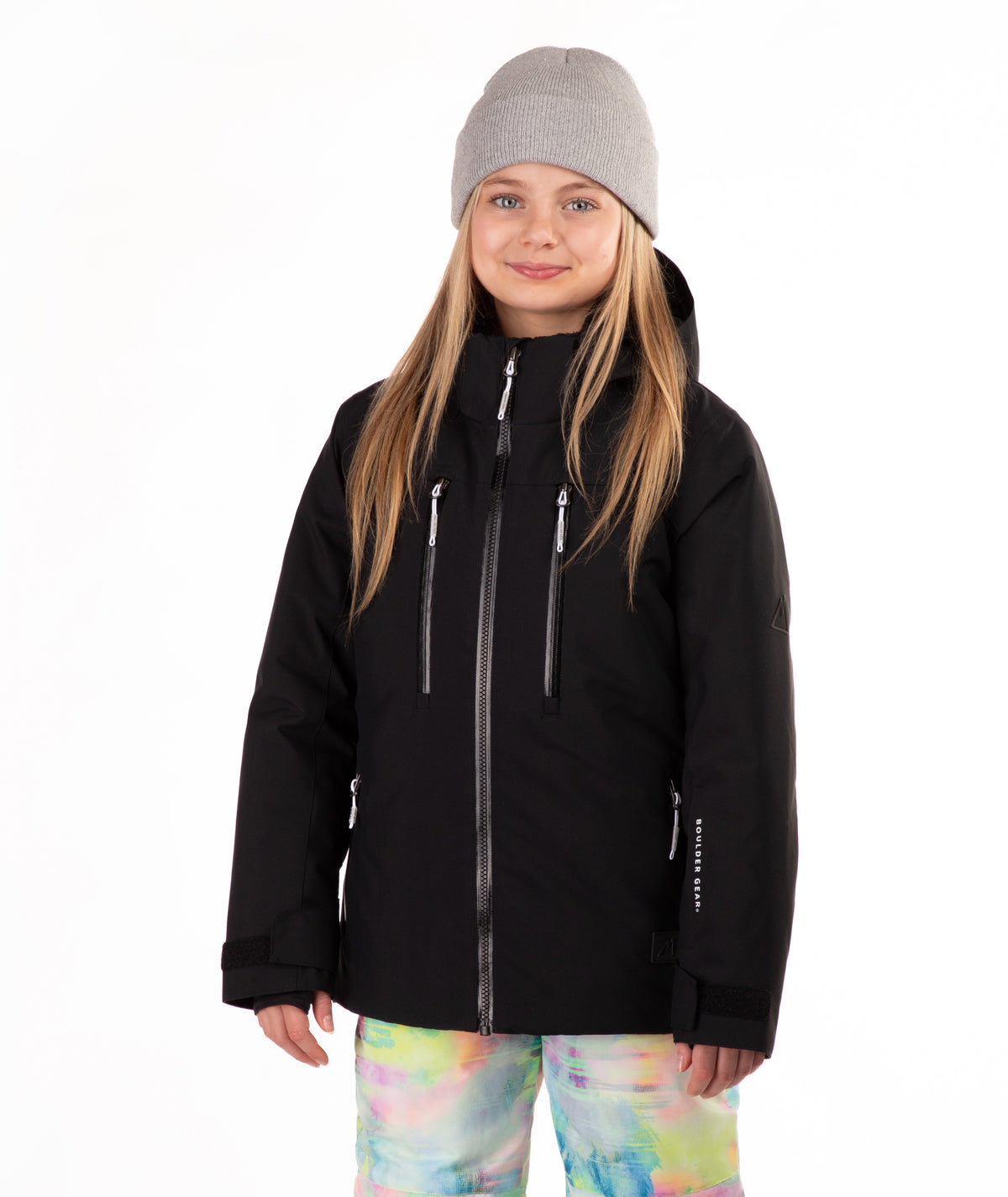 A young girl wearing a black ski jacket, a gray beanie, and colorful tie-dye snow pants, standing against a solid white background.