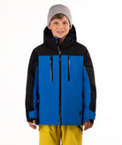 A child wearing a blue and black ski jacket, a gray beanie, and yellow ski pants, standing against a plain white studio background.