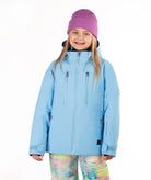 A young girl wearing a light blue ski jacket, a purple knit beanie, and colorful tie-dye snow pants, standing against a solid white background.