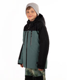 A child wearing a color-blocked ski jacket in black and muted teal, paired with a gray beanie and camouflage-patterned pants, against a plain white background.