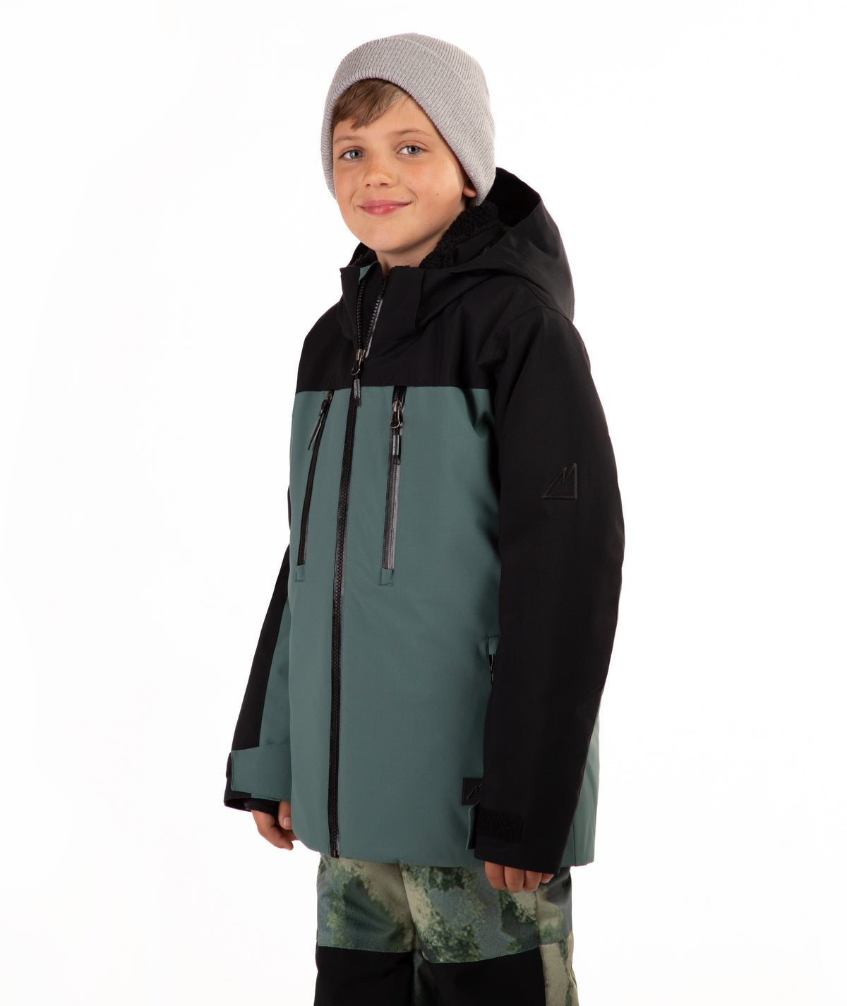 A child wearing a color-blocked ski jacket in black and muted teal, paired with a gray beanie and camouflage-patterned pants, against a plain white background.